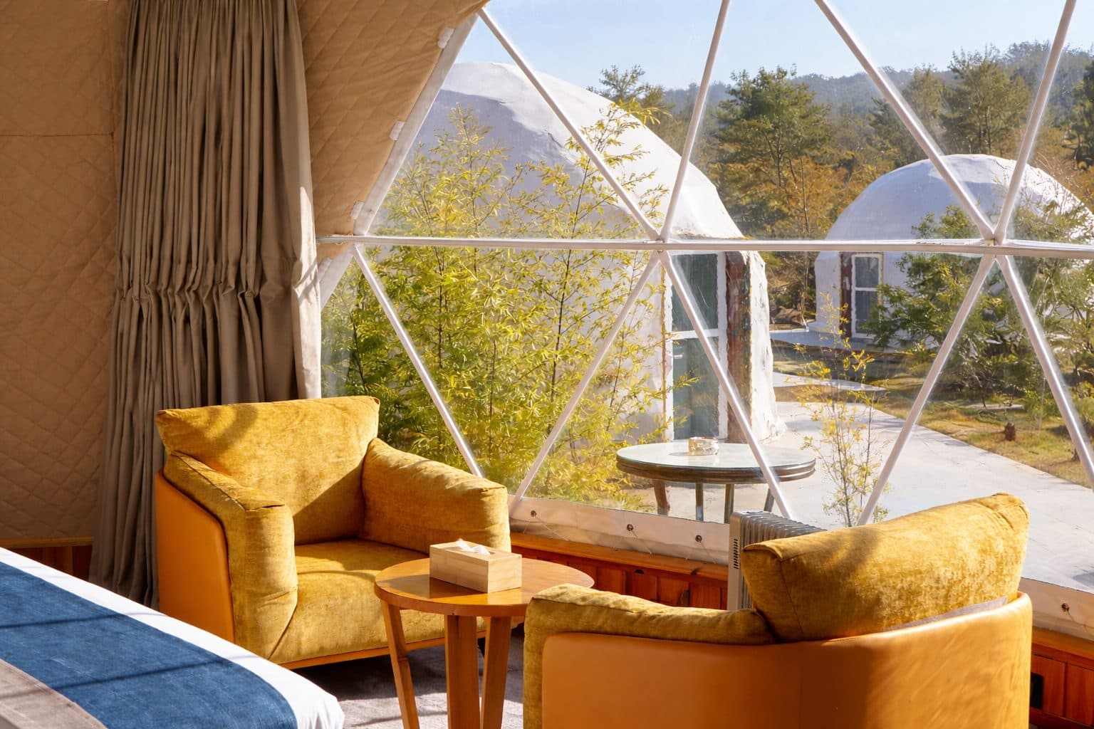 Lounge armchairs beside the panoramic geodesic window of a Deluxe Dome, looking out to the neighbouring domes and pine forest at Kepdinkha, Paro
